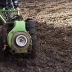 Agricultor opera um motocultivador em um campo recém-arado, preparando o solo para o plantio.
