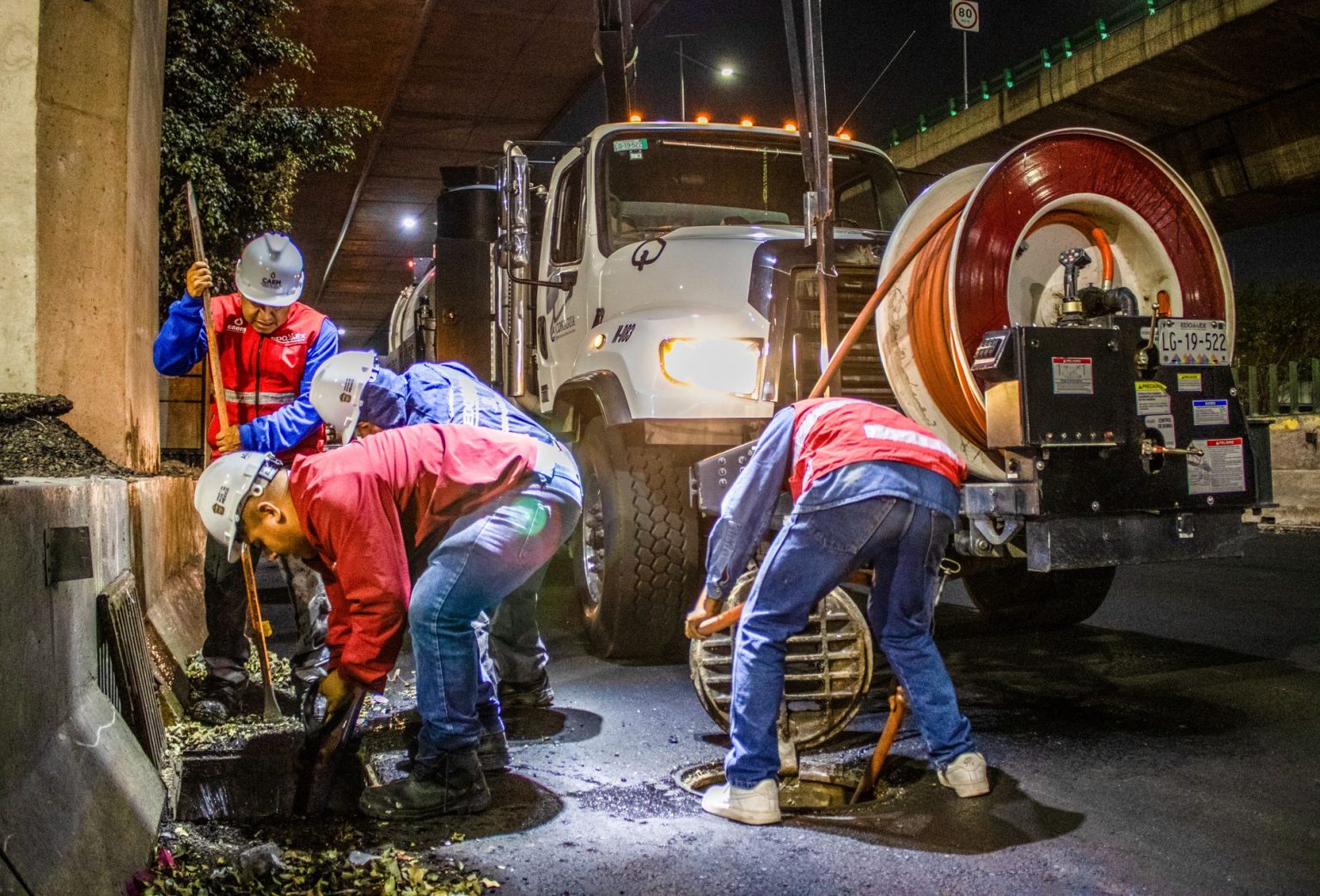 Trabalhadores realizam a limpeza de um sistema de drenagem urbana com máquinas, à noite, sob um viaduto.