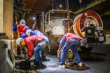 Trabalhadores realizam a limpeza de um sistema de drenagem urbana com máquinas, à noite, sob um viaduto.