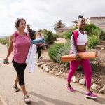Grupo de mulheres caminha ao ar livre carregando colchonetes para uma atividade física.