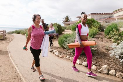Grupo de mulheres caminha ao ar livre carregando colchonetes para uma atividade física.