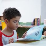 Menino lê atentamente um livro em uma sala de aula.