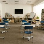 Interior de uma sala de aula moderna com estantes de livros, mesas, cadeiras e televisão.