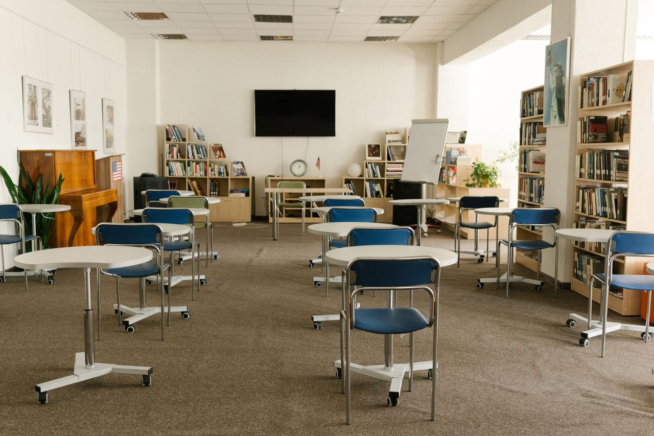 Interior de uma sala de aula moderna com estantes de livros, mesas, cadeiras e televisão.
